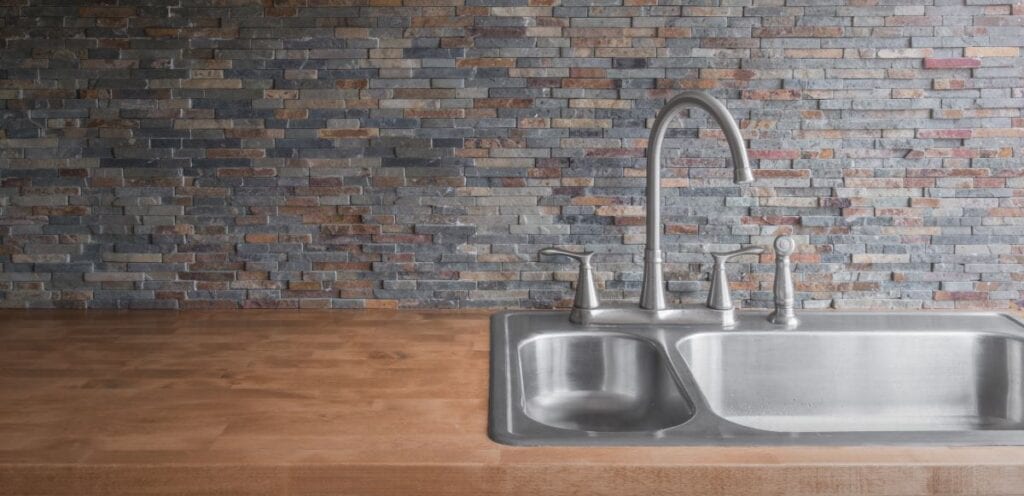 Close up of stone veneer kitchen backsplash with wood countertops and stainless steel faucet